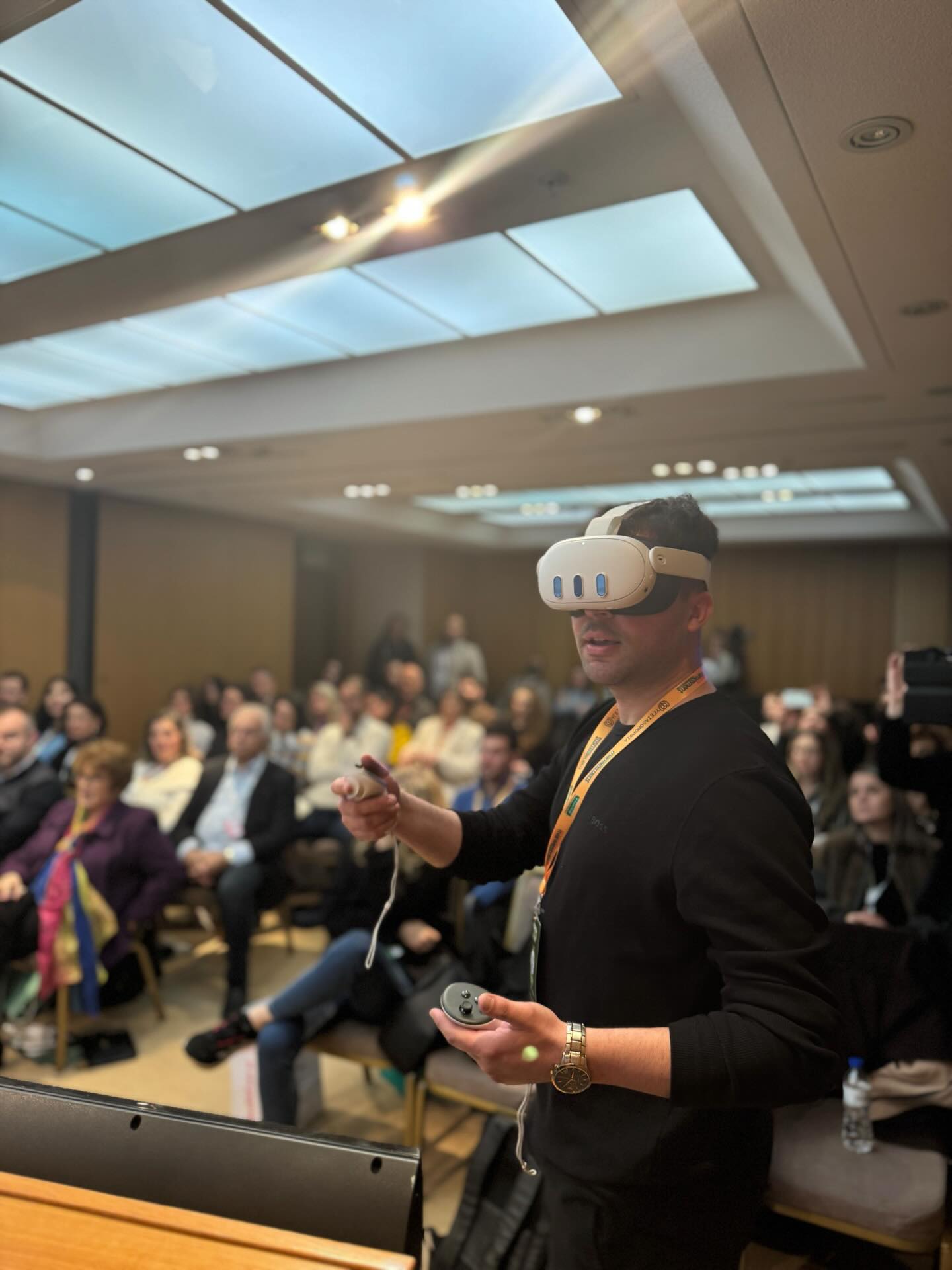 Man wearing a white VR headset and black shirt speaks to an audience at a conference holding a controller and microphone in a crowded room