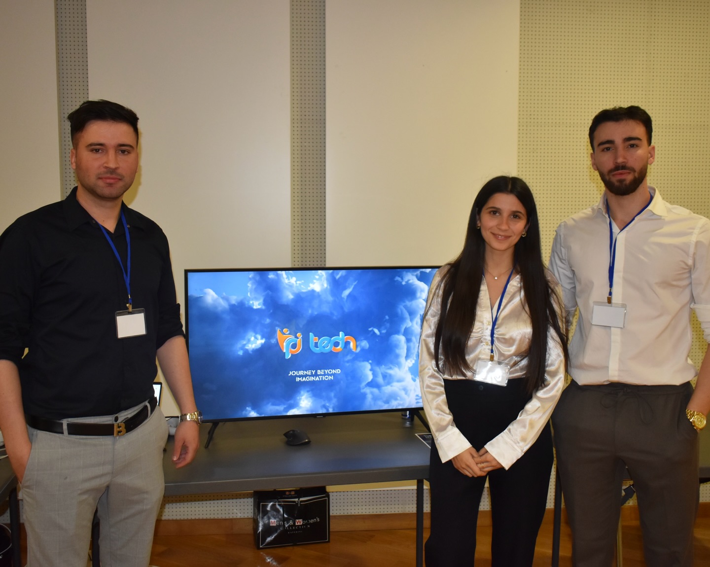 Three young professionals pose beside a monitor showing a blue sky with clouds and a colorful logo all wearing conference lanyards with badges