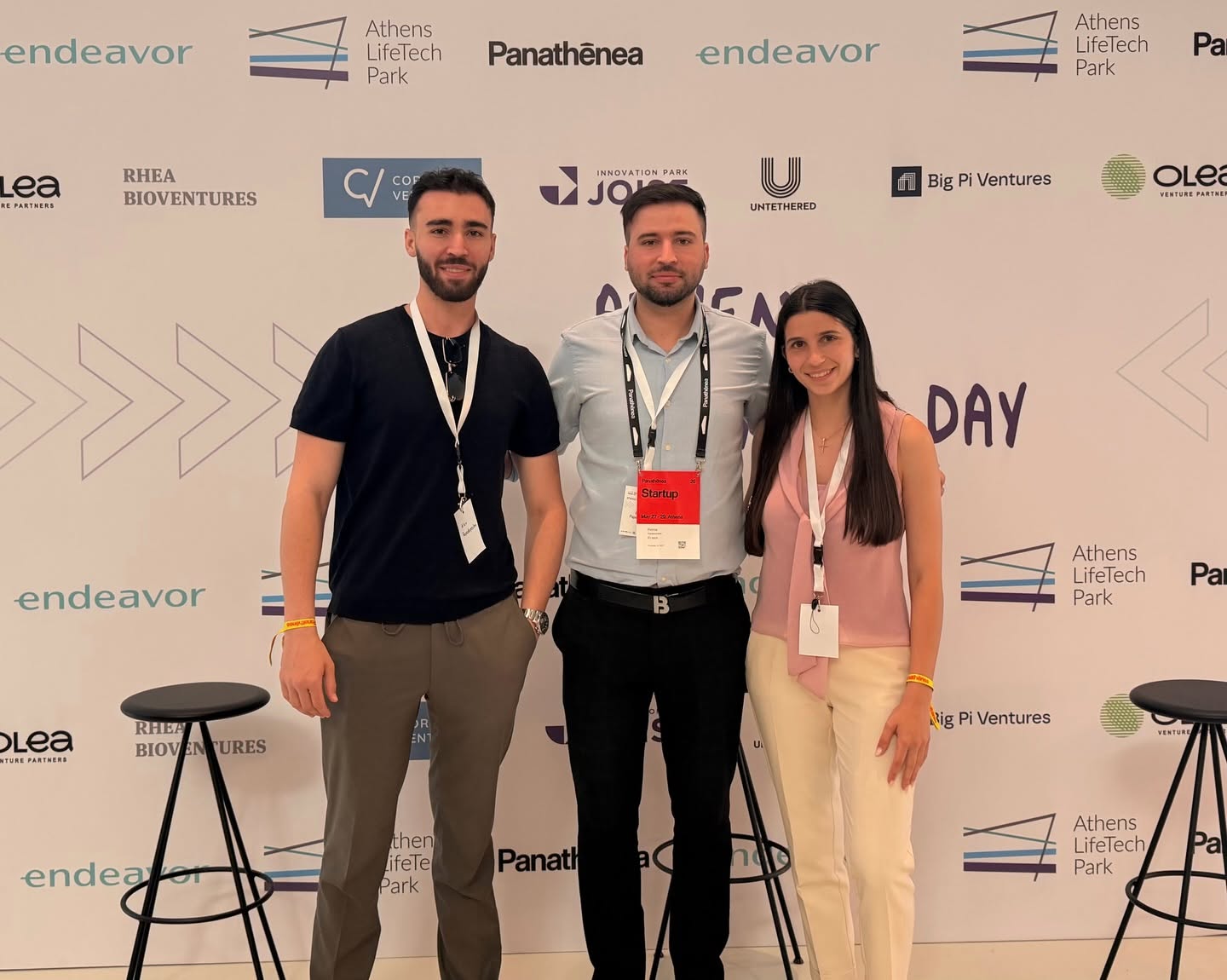 Three people pose for a photo at a startup event standing in front of a sponsor backdrop left man in a black T shirt center man in a light blue shirt right woman in a pink sleeveless top