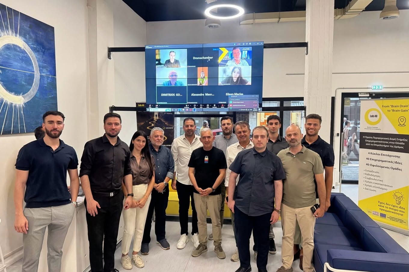 Twelve professionals pose for a group photo in a modern office lobby with a large wall mounted screen showing a Zoom call behind them