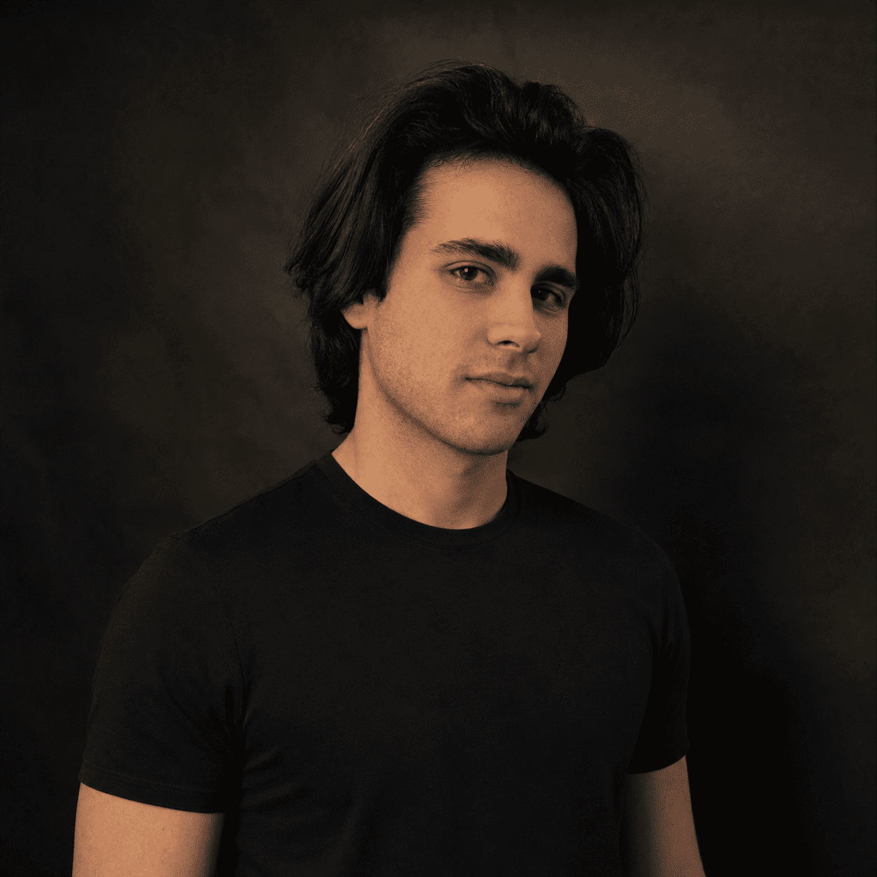 Portrait of a young man with dark hair in a black t-shirt against a dark studio background, looking slightly to the side.