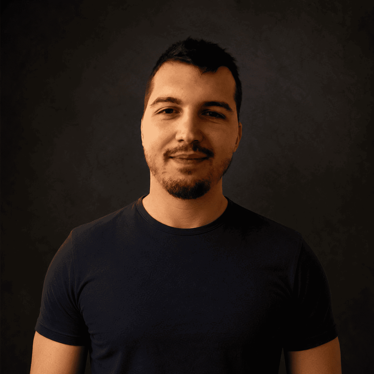 Portrait of a man with short dark hair and light beard wearing a black T-shirt, against a dark textured background.