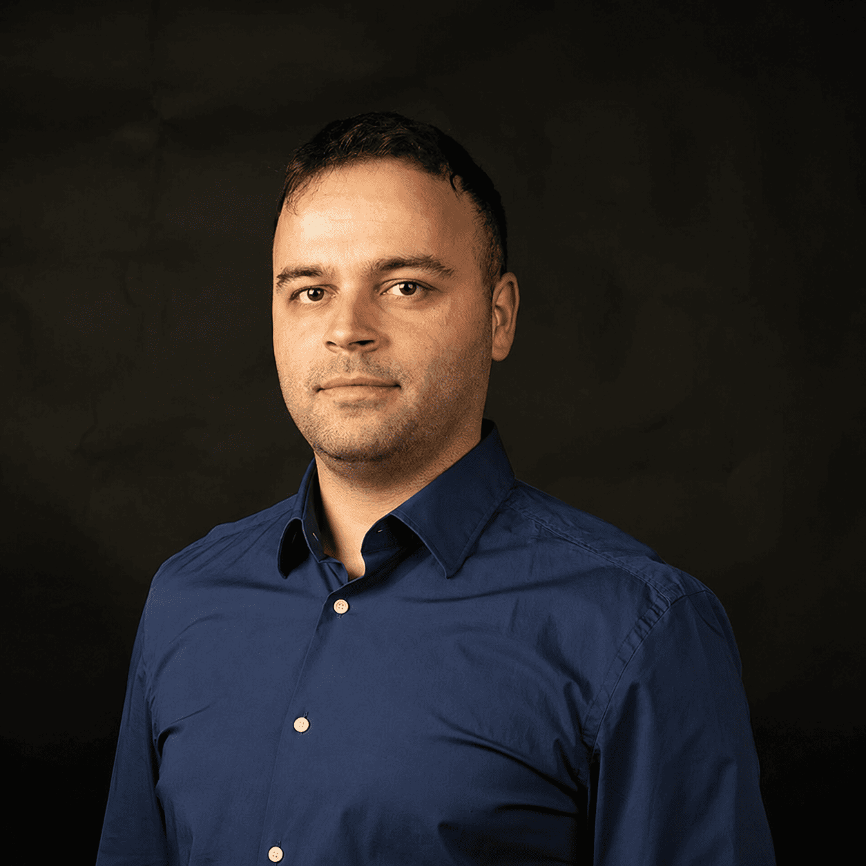 Professional headshot of a man in a blue button-down shirt against a dark background, neutral expression.